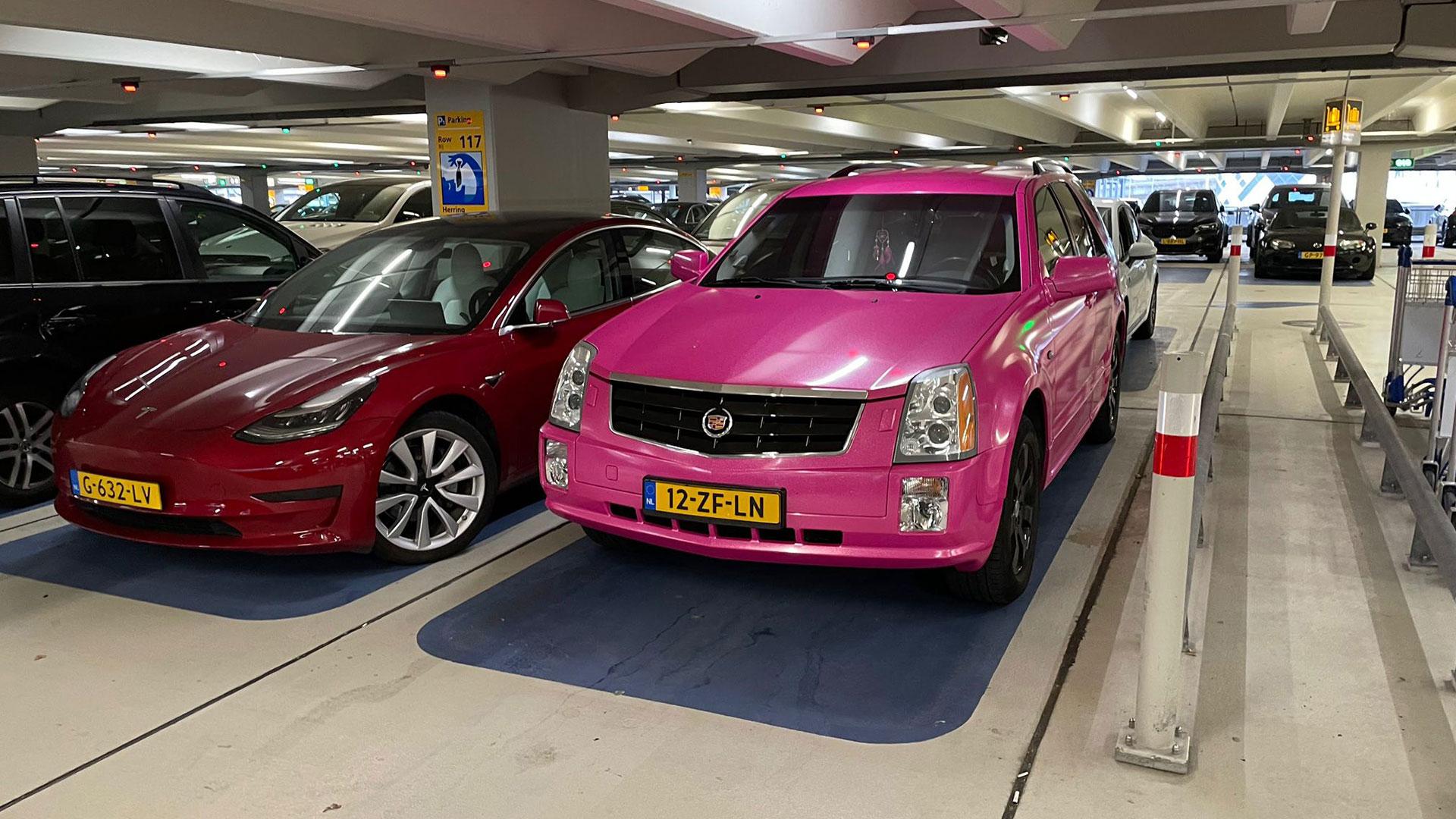 Roze Cadillac SRX geparkeerd schuin voor naast Tesla Model 3 bij Schiphol