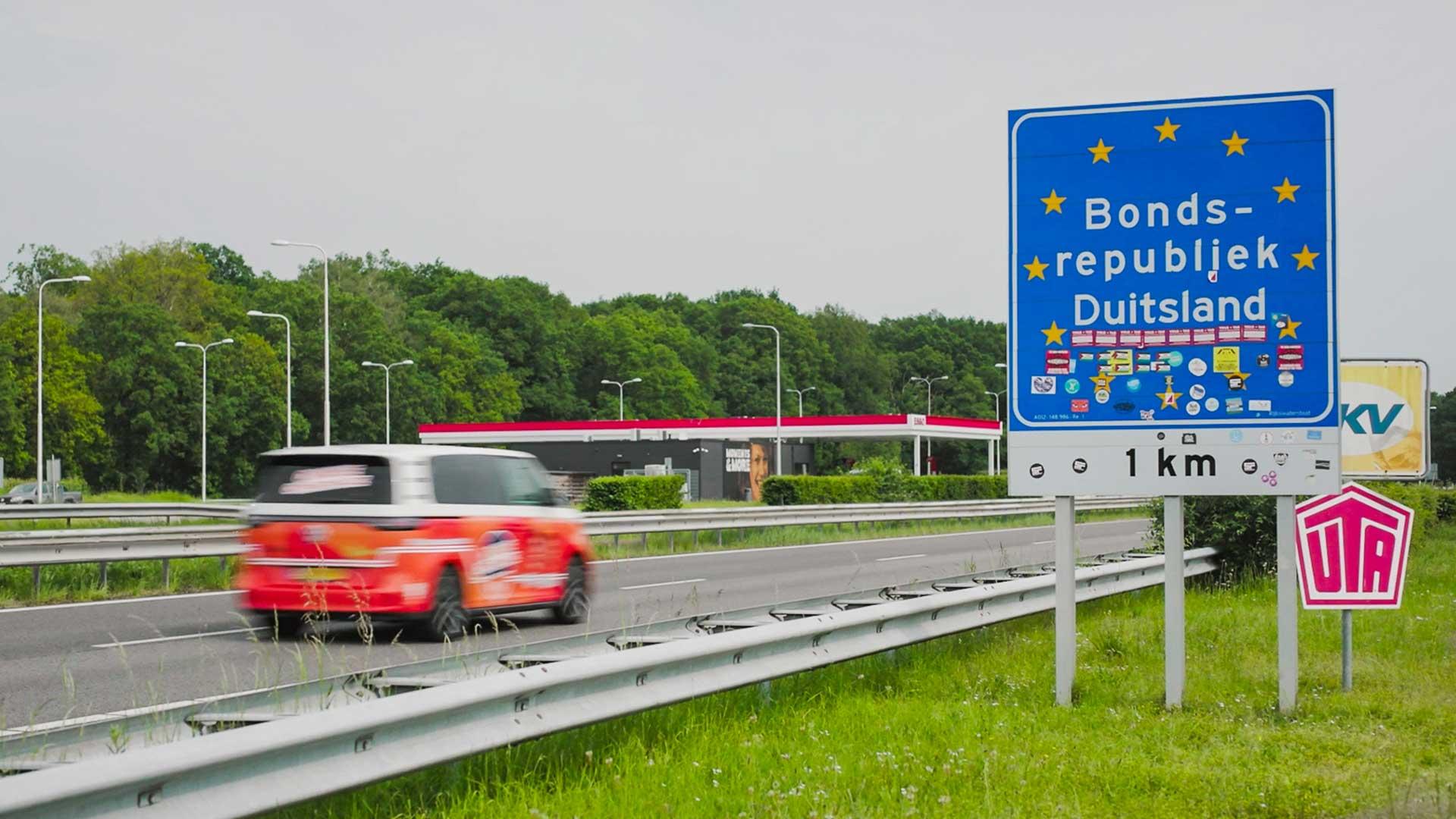 Oranje VW ID. Buzz rijdt Duitsland binnen bord Bondsrepubliek Duitsland landsgrens grens
