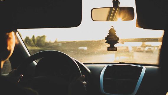 Bestuurder rijdend op een weg met zon tegen zonsondergang met boompje luchtverfrisser aan de binnenspiegel (Unsplash)